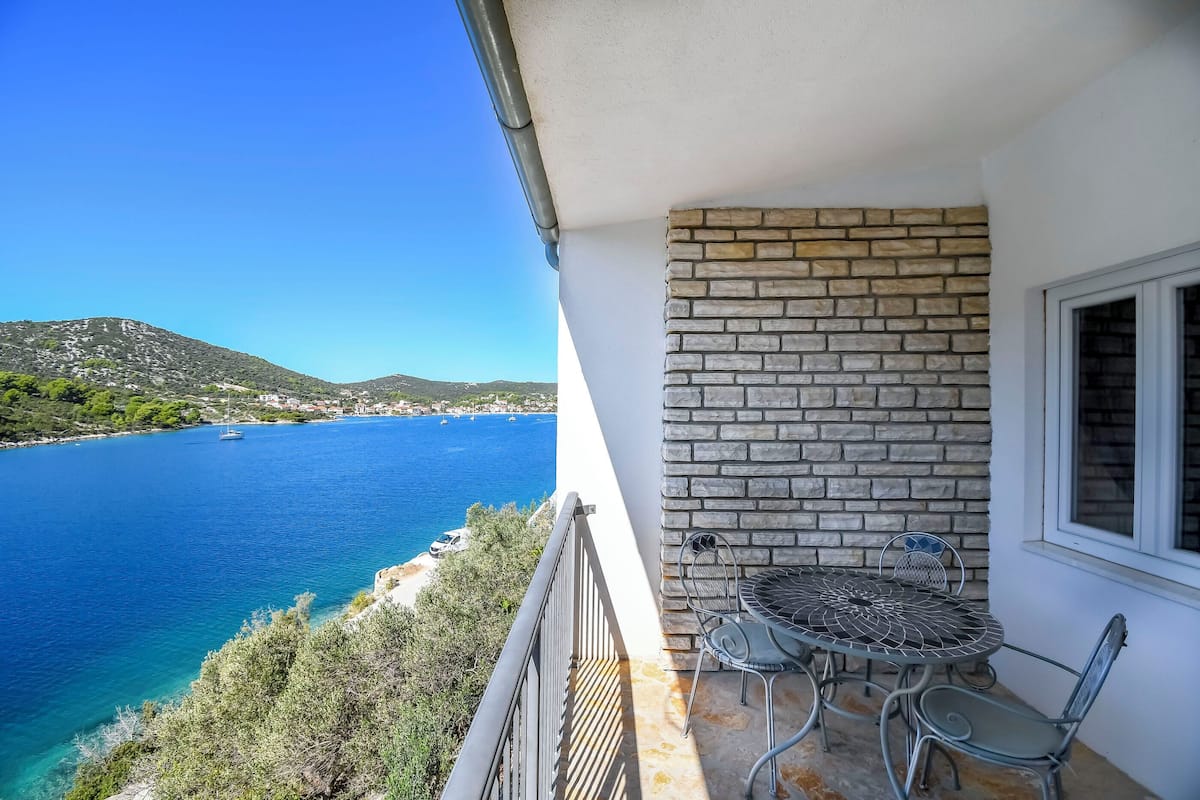 Ein Balkon mit Steinmauer, einem runden Tisch mit Stühlen und Blick auf eine Küstenlandschaft mit Booten.