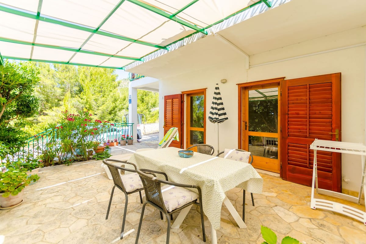 A covered outdoor patio with a table and chairs, surrounded by potted plants and a view of the garden.