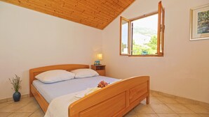 Een slaapkamer met een houten bed, een nachtkastje, een bedlampje, een raam met een open luik en een kamerplant.