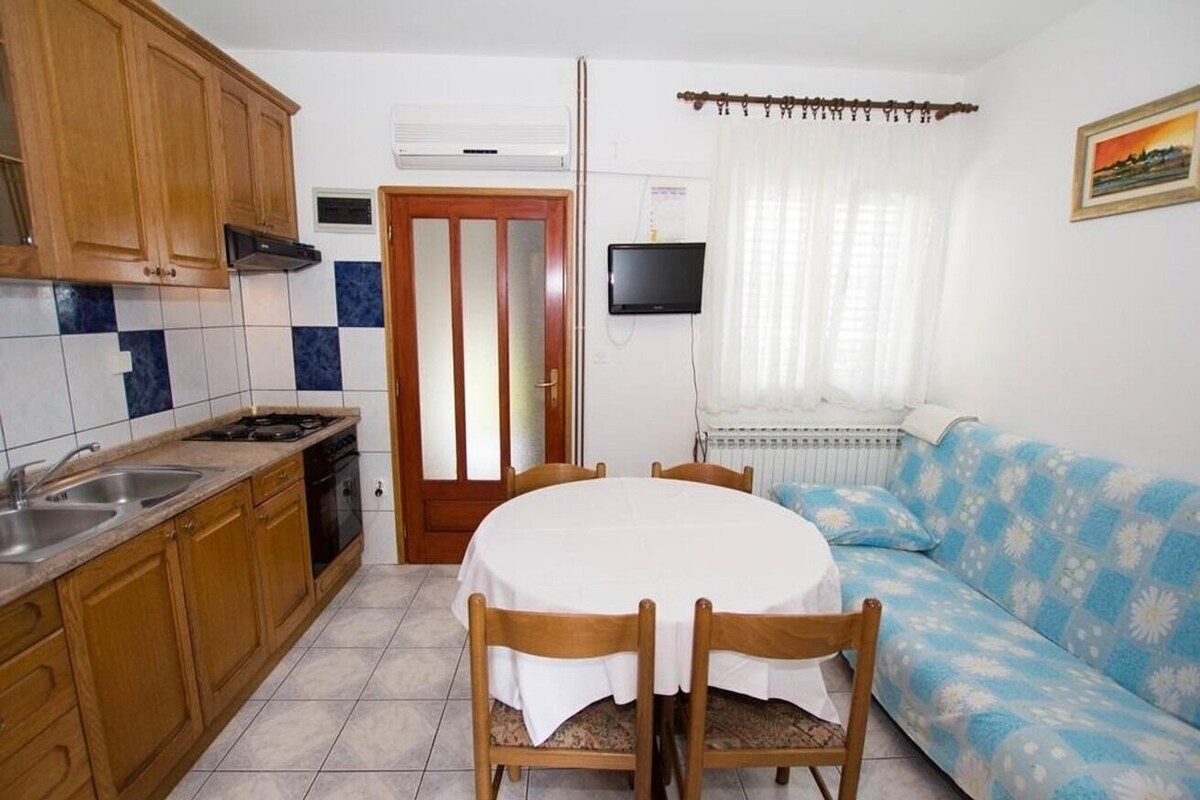 A compact kitchen with wooden cabinets, a white tiled backsplash, a dining table with chairs, a sofa, and a television mounted on the wall.