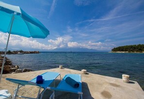 Eine Strandkulisse mit einer Betonsteg, zwei blauen Liegestühlen, einem blauen Sonnenschirm und freiem Blick auf das Meer und das entfernte Land.
