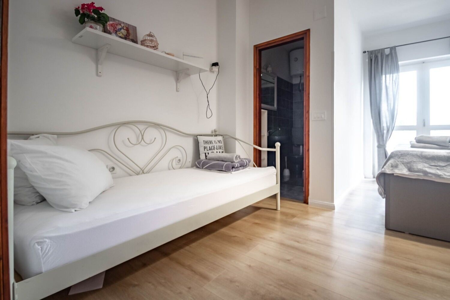 Une chambre moderne avec un canapé-lit blanc, un plancher en bois, une petite étagère ornée d’objets décoratifs et une vue sur la salle de bain par une porte ouverte.