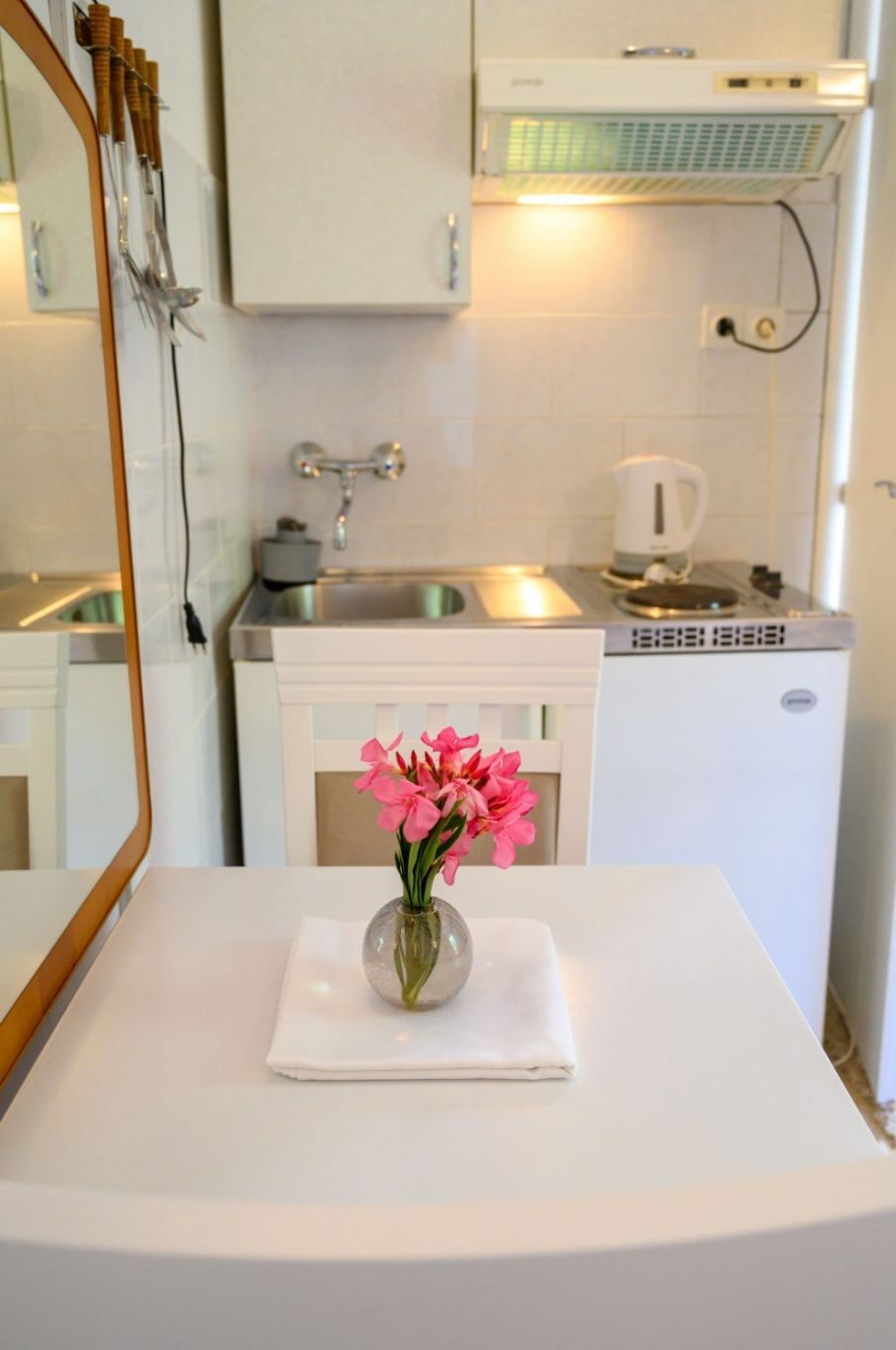 Et lille hvidt bord med en vase fyldt med lyserøde blomster, en hvid serviet og et spejl.