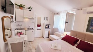 A compact hotel room with a kitchenette, a bed with red and white pillows, a bedside table, and a large mirror. - Apartments Goge (Gradac)