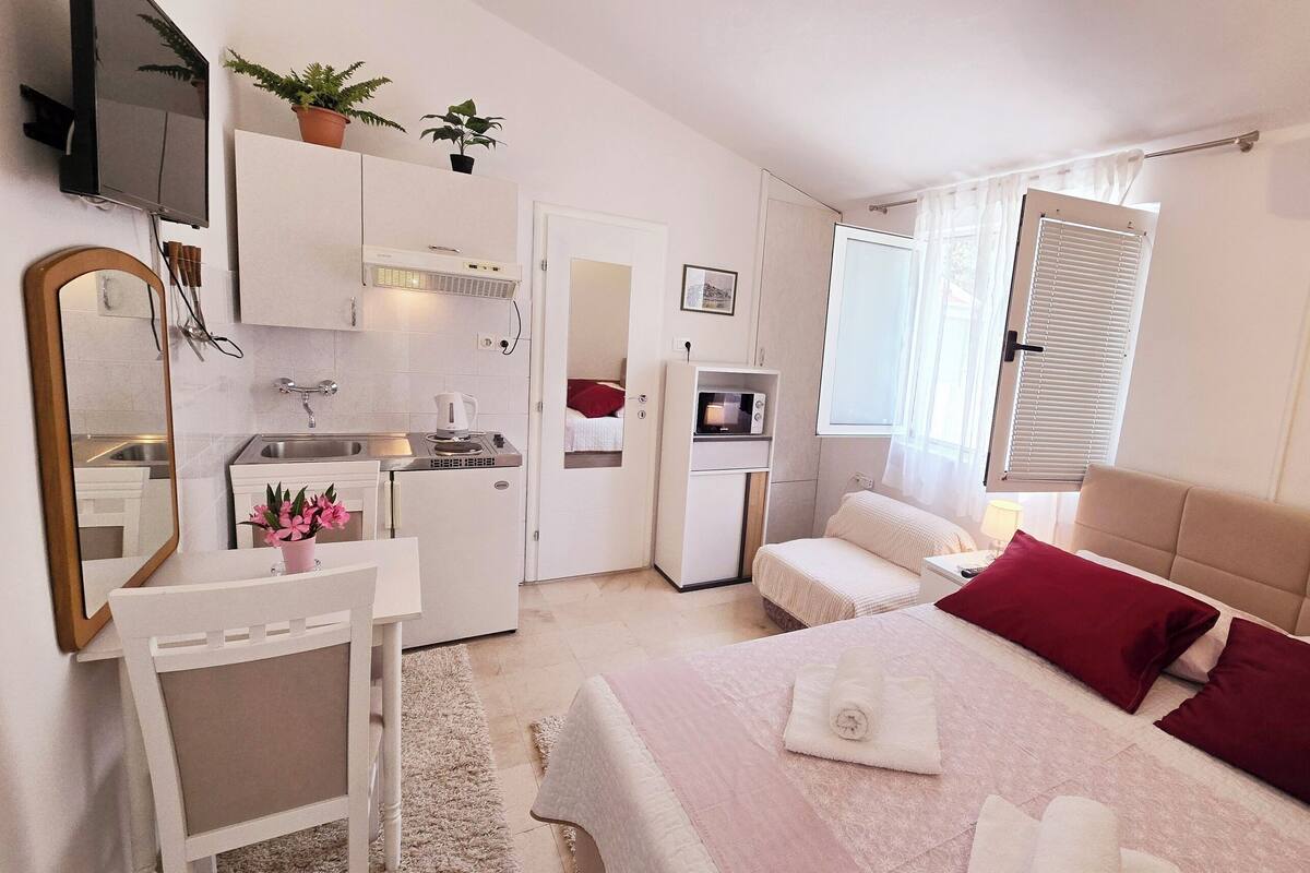 A compact hotel room with a kitchenette, a bed with red and white pillows, a bedside table, and a large mirror.