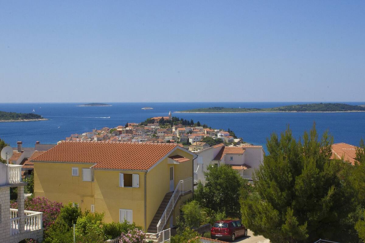 Une ville côtière aux bâtiments aux toits de tuiles rouges, bordée par une mer d’un bleu limpide, avec une île qui se profile à l’horizon.