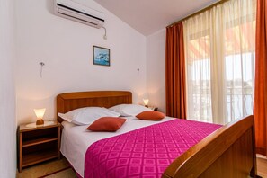 A hotel room with a bed, a nightstand, an air conditioner, a window with curtains, and a wall-mounted picture. - Apartment VV (Seget)