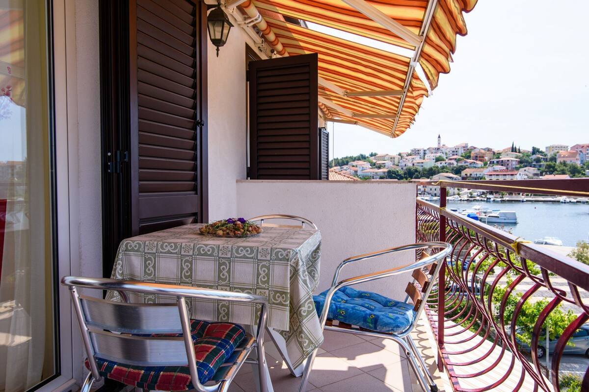 A balcony with a table and chairs, a view of a town and water, and a railing.
