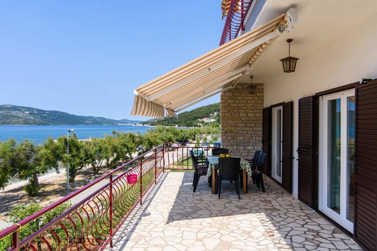 A balcony with a stone floor, a table with chairs, and a view of the sea and hills.