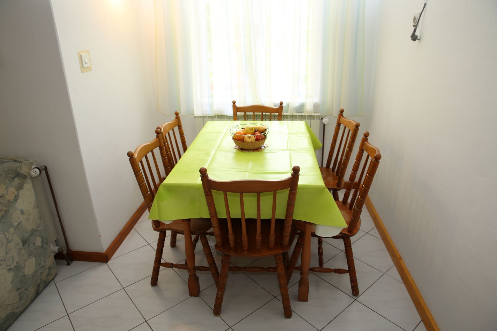 Una mesa de comedor con mantel verde, rodeada de sillas de madera, en una habitación con paredes blancas y un radiador.
