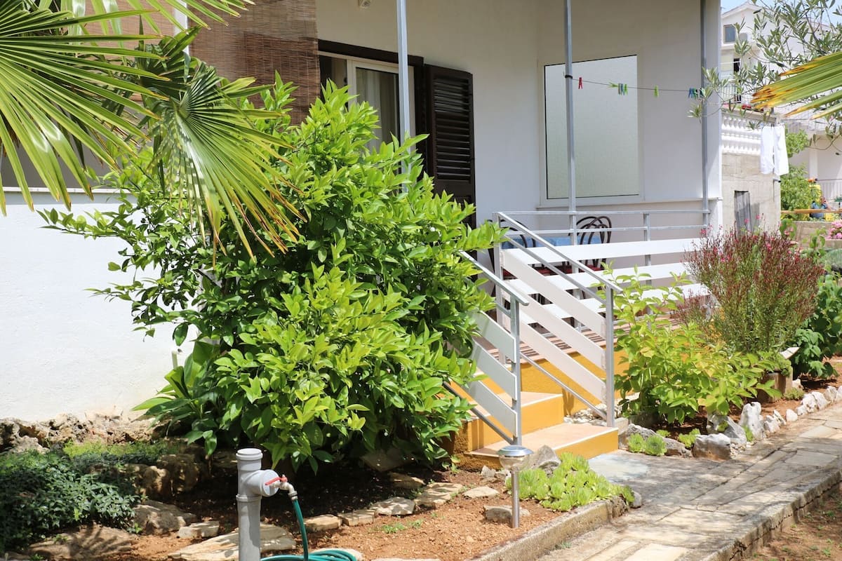 Un immeuble résidentiel à la façade blanche, doté d’un escalier gris et d’un espace jardin agrémenté de diverses plantes et d’un élément décoratif aquatique.