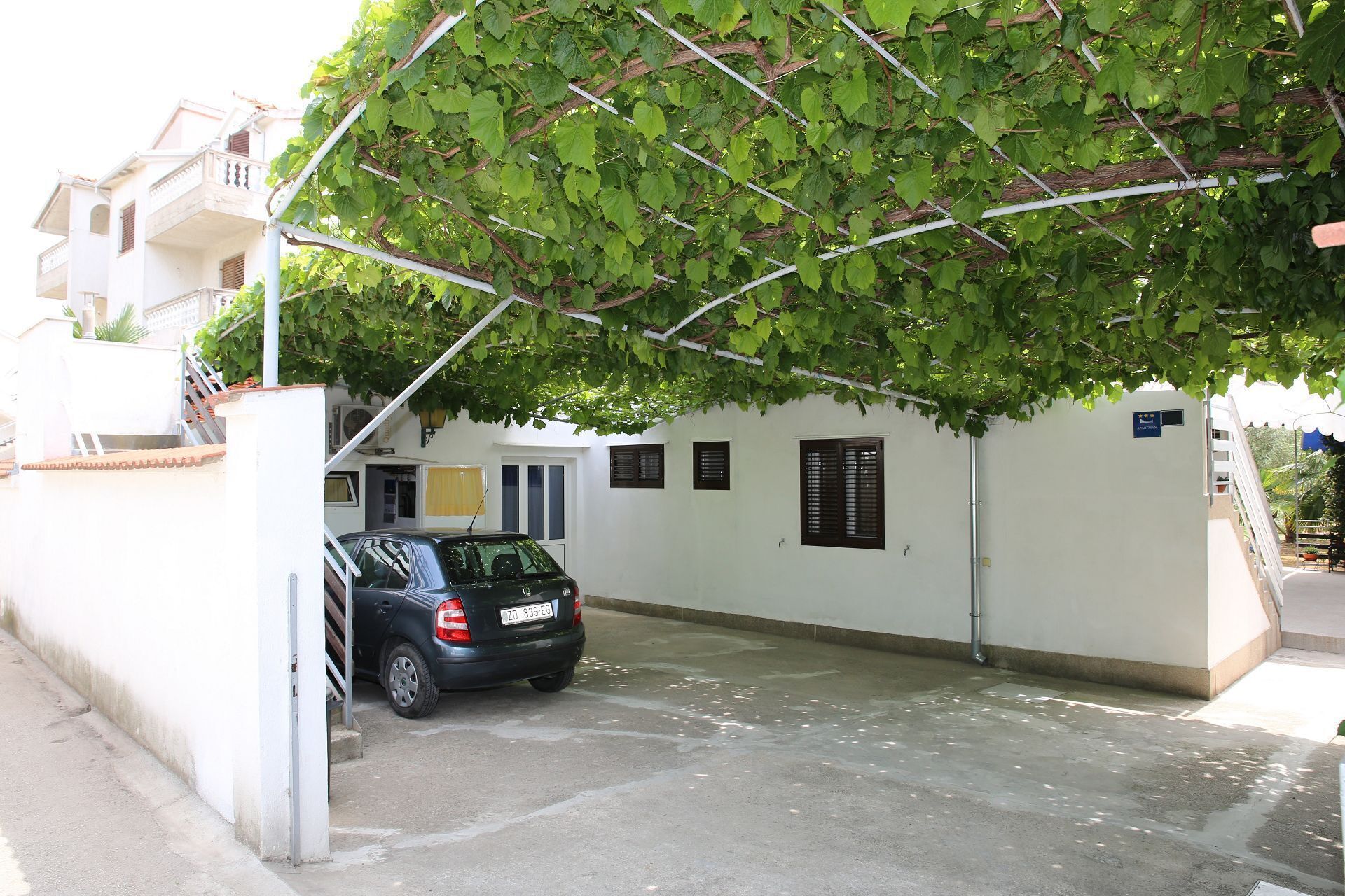 A car parked in a covered parking area with a white building and a balcony in the background.