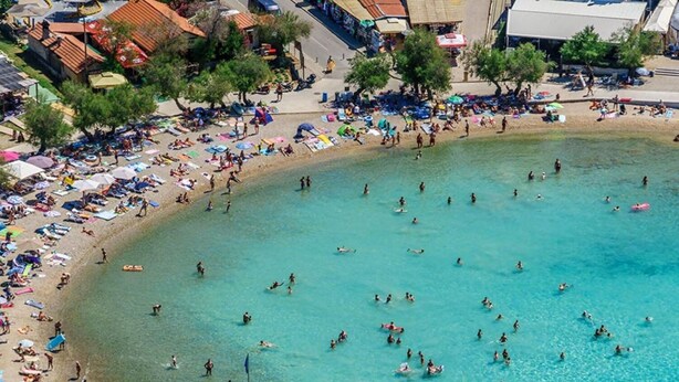 Eine Luftaufnahme eines Strandes mit klarem, türkisblauem Wasser, einem Steg mit Sonnenschirmen sowie Menschen, die sonnenbaden und schwimmen.