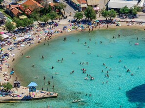 An aerial view of a beach with clear turquoise water, a pier with umbrellas, and people sunbathing and swimming. - Apartments Araz (Vodice)