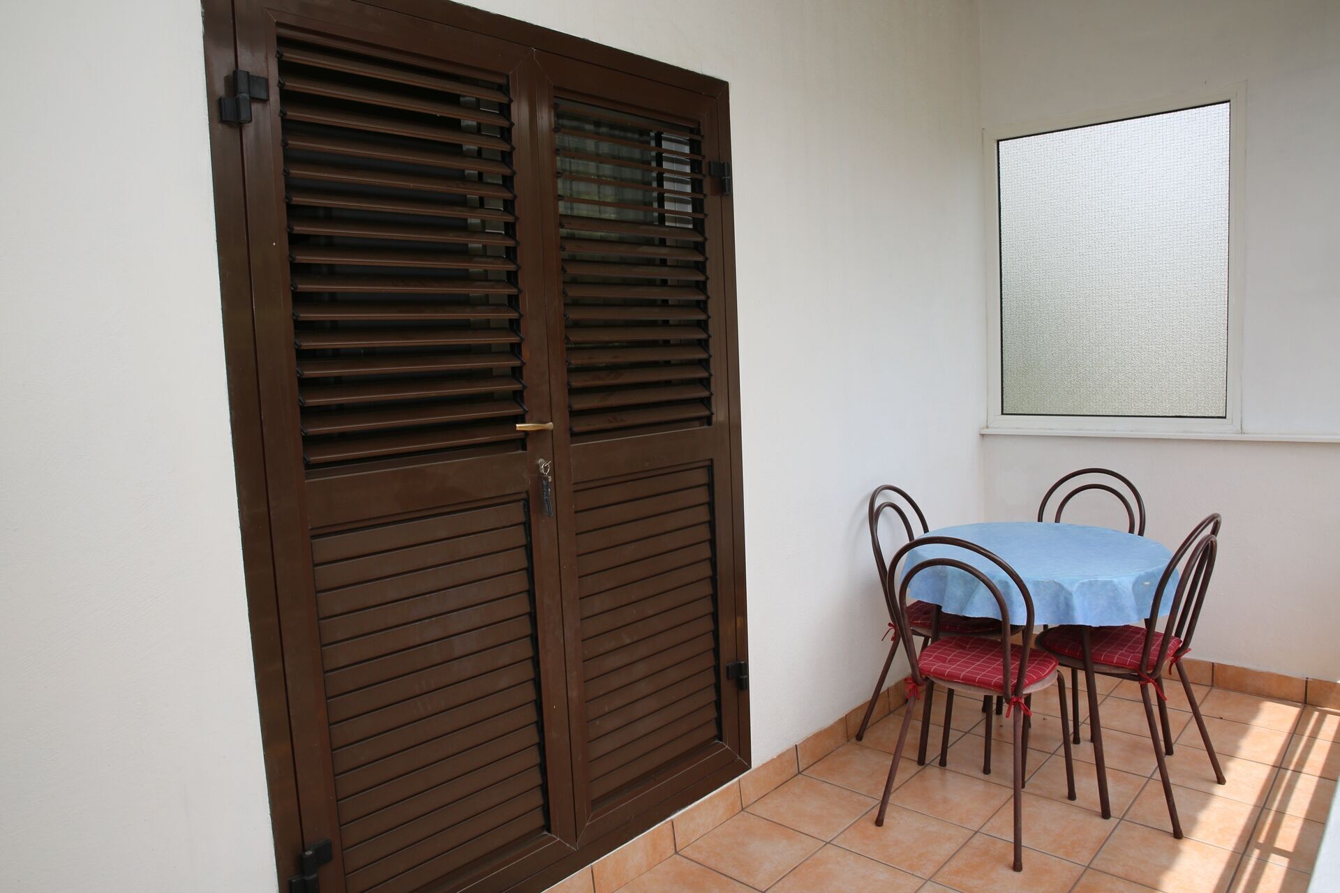 Una habitación con una puerta marrón cerrada, una ventana con vidrio esmerilado, una mesa azul con cuatro sillas y un piso de baldosas.