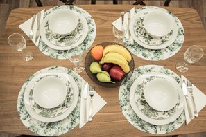 A dining table set with floral-patterned plates, bowls, and cutlery, accompanied by a fruit basket and wine glasses. - Socrates - Premium Athenian Apartment near Acropolis (Athina)