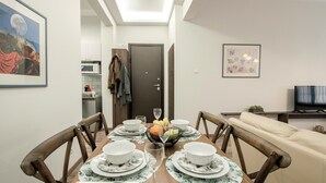 Un coin repas avec une table en bois dressée pour quatre personnes, entourée de chaises. On aperçoit un saladier de fruits sur la table, et un téléviseur est visible au fond.