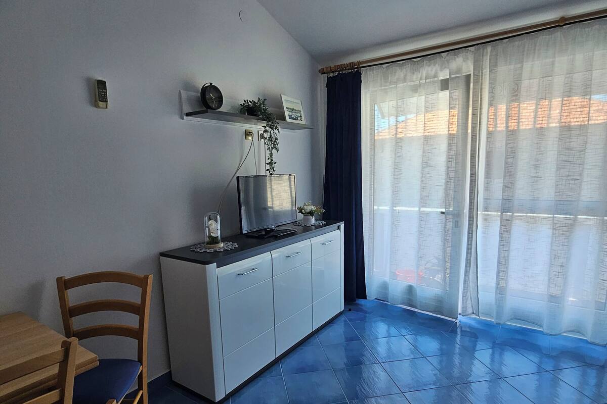 A room with a dining table, chairs, a TV, a cabinet, and a large window with curtains.