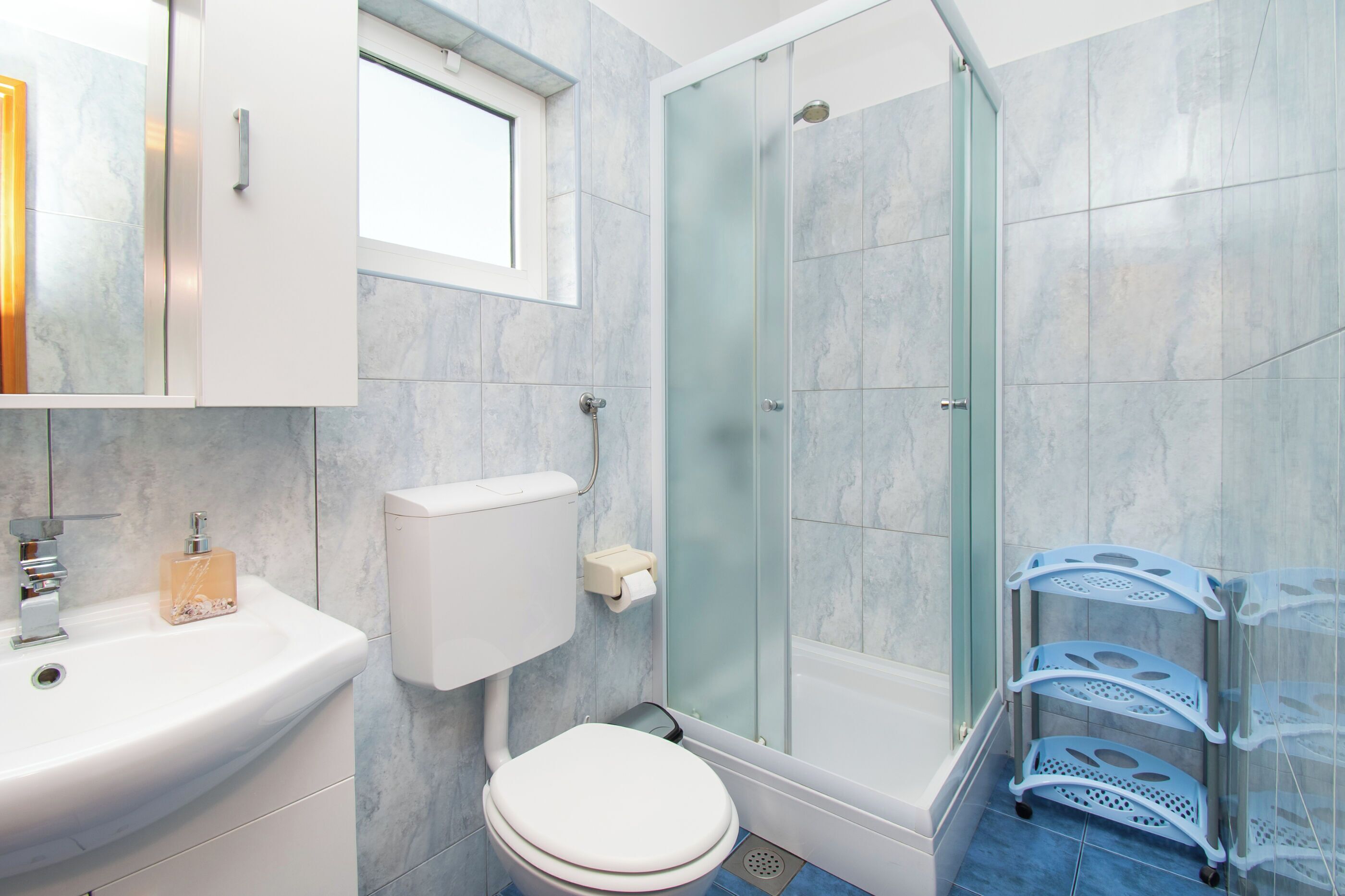 Un baño con inodoro blanco, ducha con cerramiento de vidrio, lavamanos blanco y un carro de almacenamiento azul.