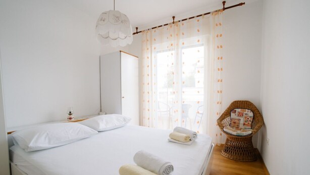A neatly made bed with two pillows and two rolled towels, a wicker chair, and a large window with curtains. - Apartments Petri (Tisno)
