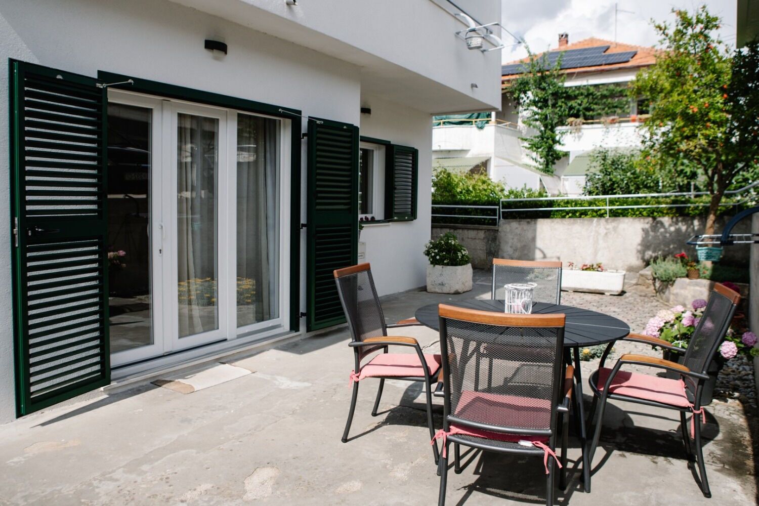 Patio esterno con tavolo rotondo e quattro sedie, una porta scorrevole in vetro e una piccola zona giardino con piante in vaso.