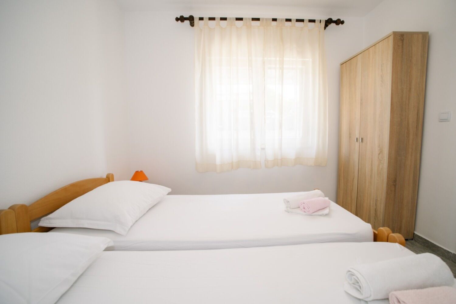 A bedroom with two single beds, a wooden wardrobe, a window with sheer curtains, and a small orange lamp.