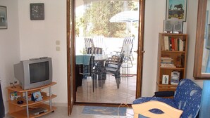 Ett vardagsrum med en tv-apparat på ett träbord, en bokhylla, ett matbord med stolar och en utsikt genom en glasdörr till ett utomhusområde med ett bord och stolar.