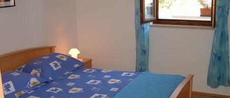Une chambre à coucher avec un lit, une lampe de chevet, une fenêtre avec des rideaux et une vue sur la verdure à l’extérieur.