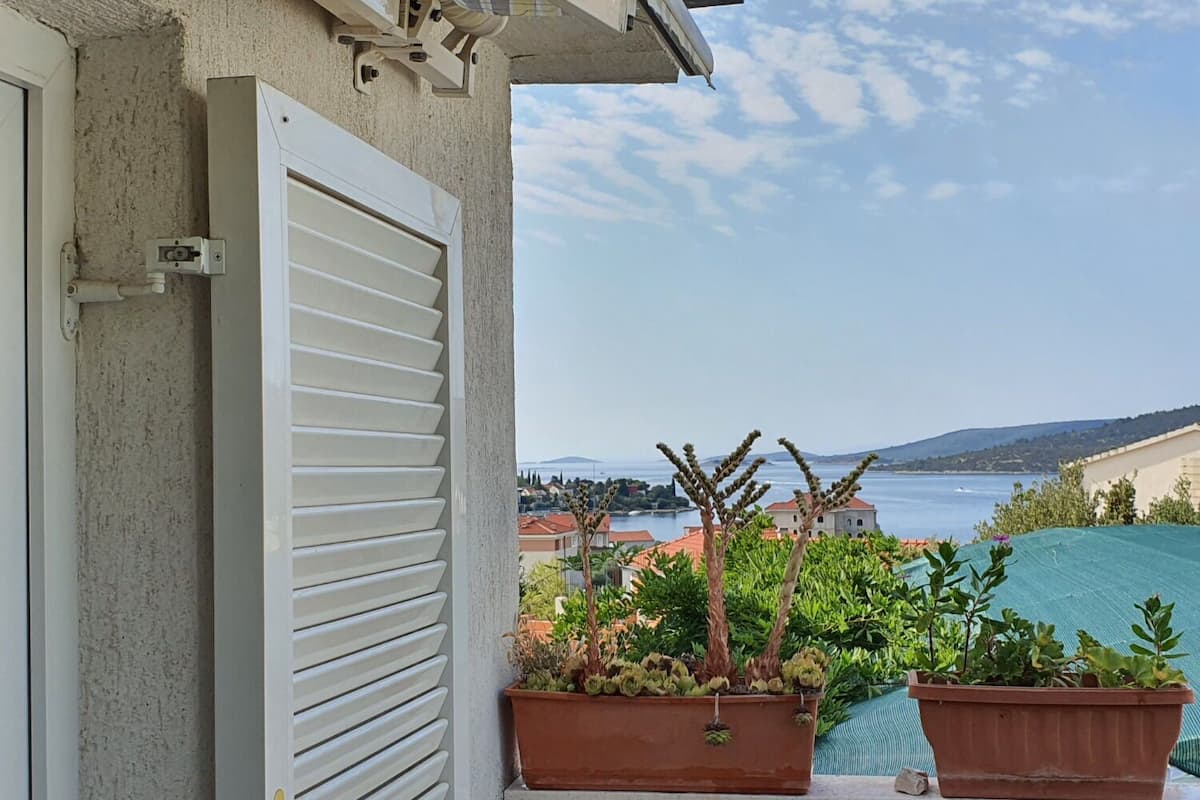 Un balcon avec des volets blancs, des plantes en pot et une vue sur un paysage côtier.