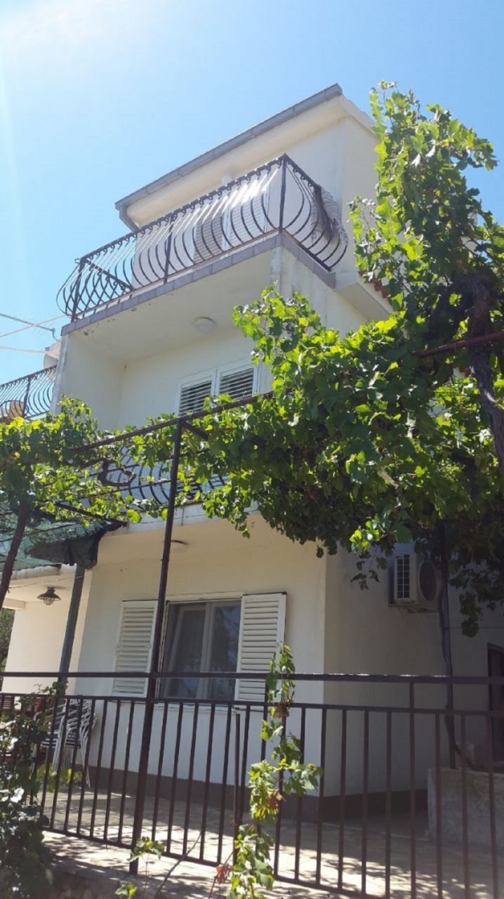Ein zweistöckiges Gebäude mit einem Balkon, einem Fenster mit Rollläden und einem Baum im Vordergrund.