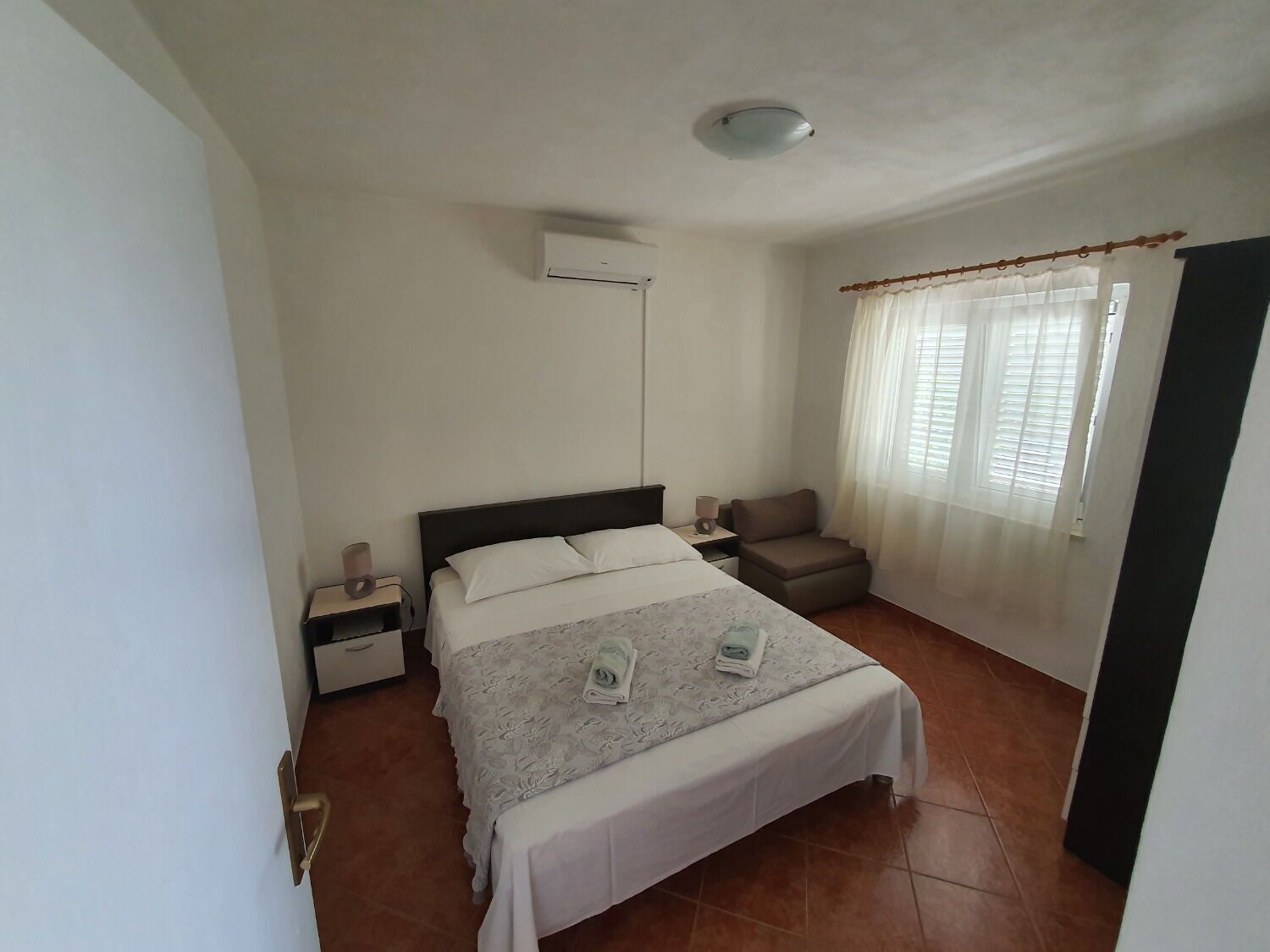 Un dormitorio con cama, mesitas de noche, una silla, ventana con cortinas y un equipo de aire acondicionado.