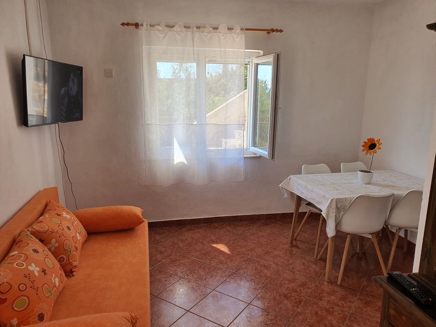 A small, bright room with a sofa, a dining table with chairs, a TV, and a window with a view of greenery.