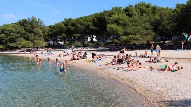 A beach with clear shallow waters, people sunbathing and swimming, and trees in the background. - Apartments Ive (Tisno)