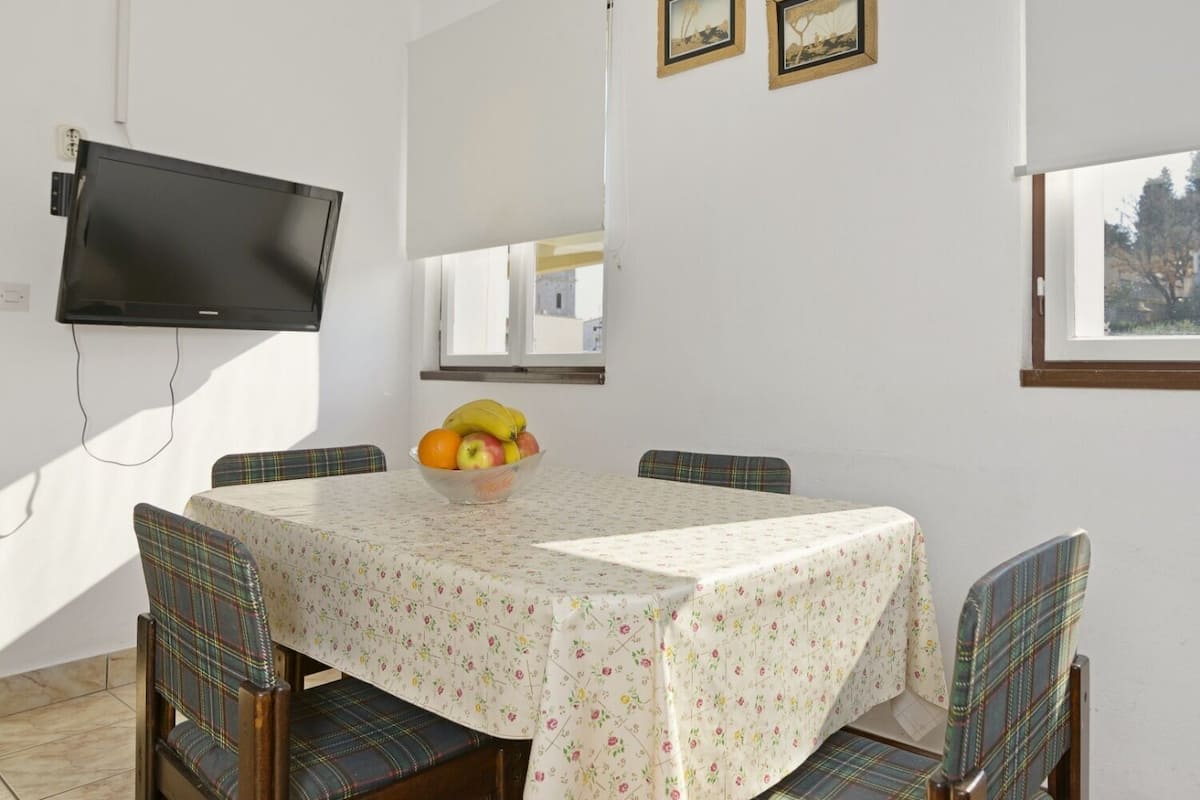 A dining area with a round table, plaid chairs, a fruit bowl, and a TV mounted on the wall.