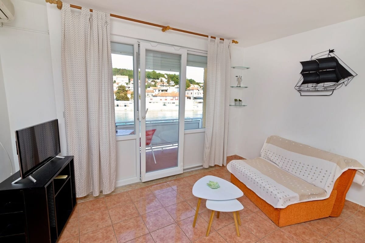 A living room with a sofa, a TV, a small table, and a balcony with a view of buildings and water.