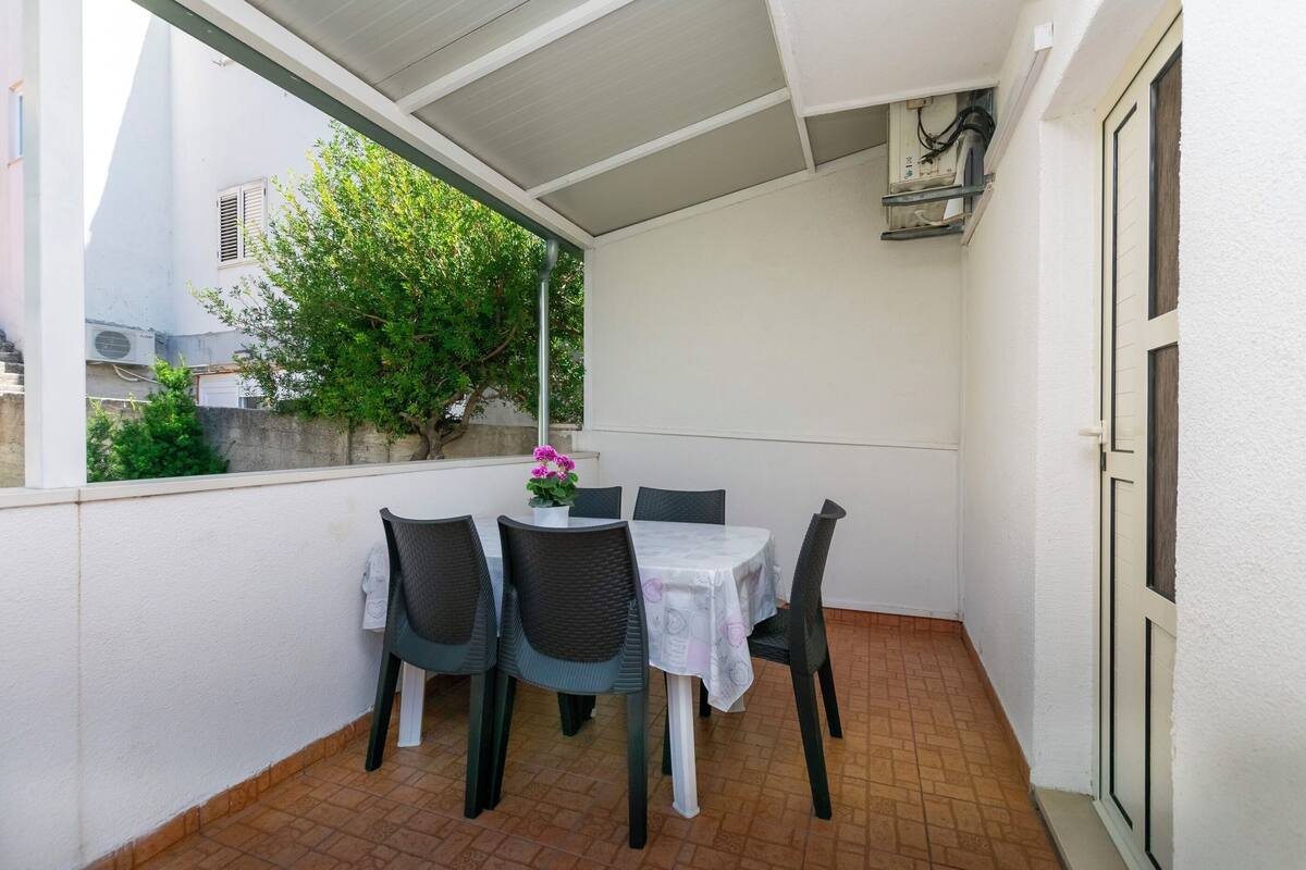 A covered balcony with a table and chairs, a potted plant, and a view of the street and greenery.