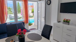 Une chambre moderne avec un balcon, une table à manger avec des sets de table, un canapé avec des coussins, un téléviseur fixé au mur et un meuble agrémenté d’un arrangement floral.