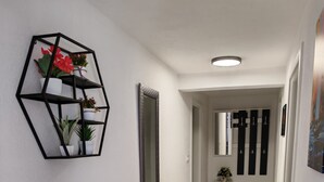 Un couloir avec une étagère murale aux motifs géométriques, sur laquelle sont disposées des plantes, un miroir et une porte qui donne sur une pièce contenant un lit et une commode.