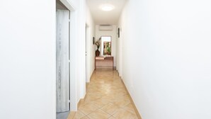 A narrow hallway with a tiled floor, a wooden door on the left, and a desk with a vase and picture frames in the background. - Apartment Bogomir (Podgora)