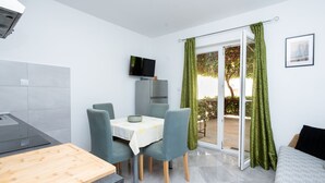 A modern kitchen with a built-in stove, a dining area with a table and chairs, a TV mounted on the wall, and a sliding glass door leading to an outdoor area. - Apartment Bogomir (Podgora)