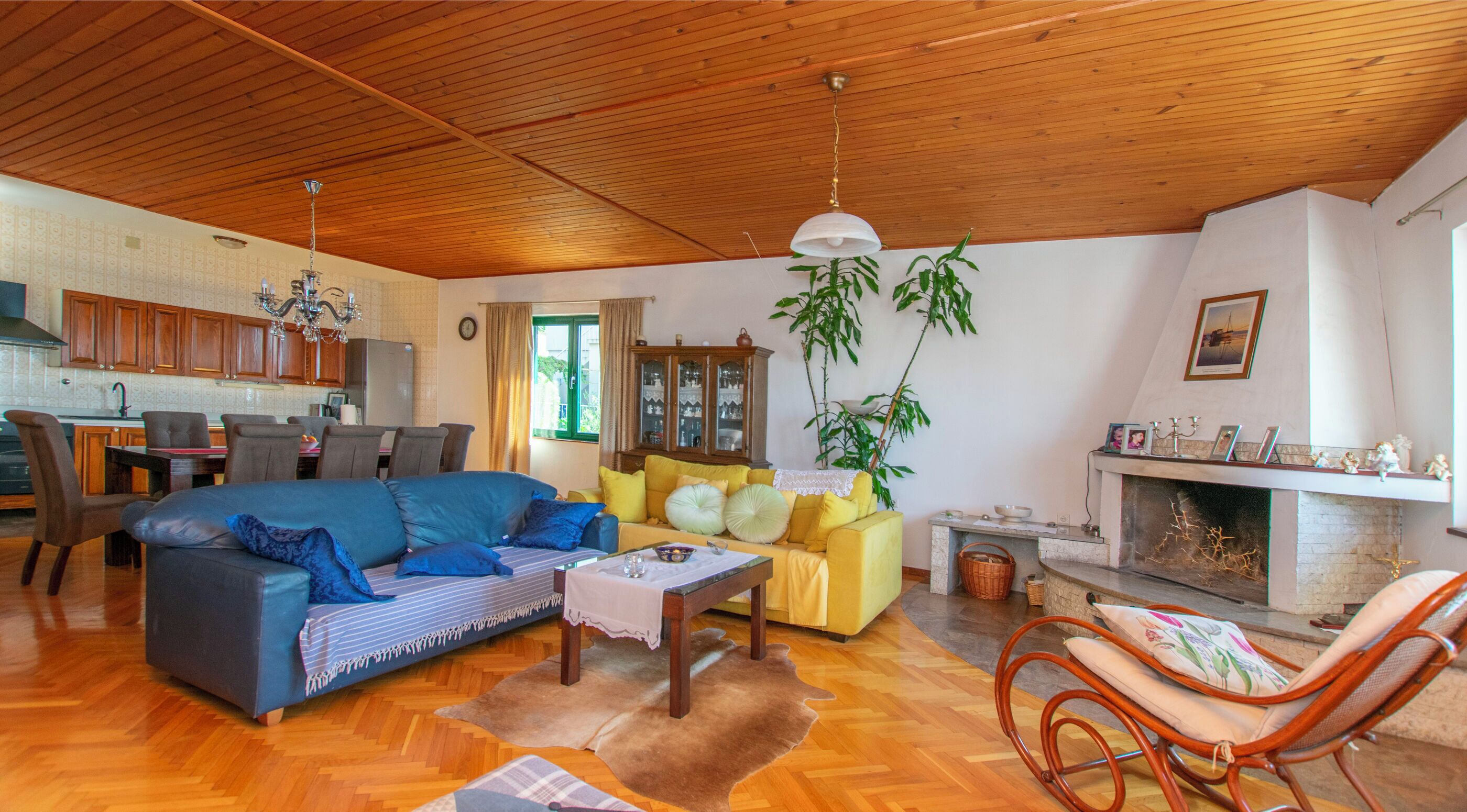 A spacious living room with a wooden ceiling, a dining area with a kitchen in the background, and a fireplace with decorative items.