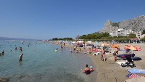 Una spiaggia affollata con persone che nuotano e prendono il sole, ombrelloni e una collina rocciosa sullo sfondo.