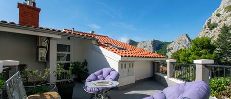 A terrace with a red-tiled roof, outdoor seating, and a view of rocky mountains.
