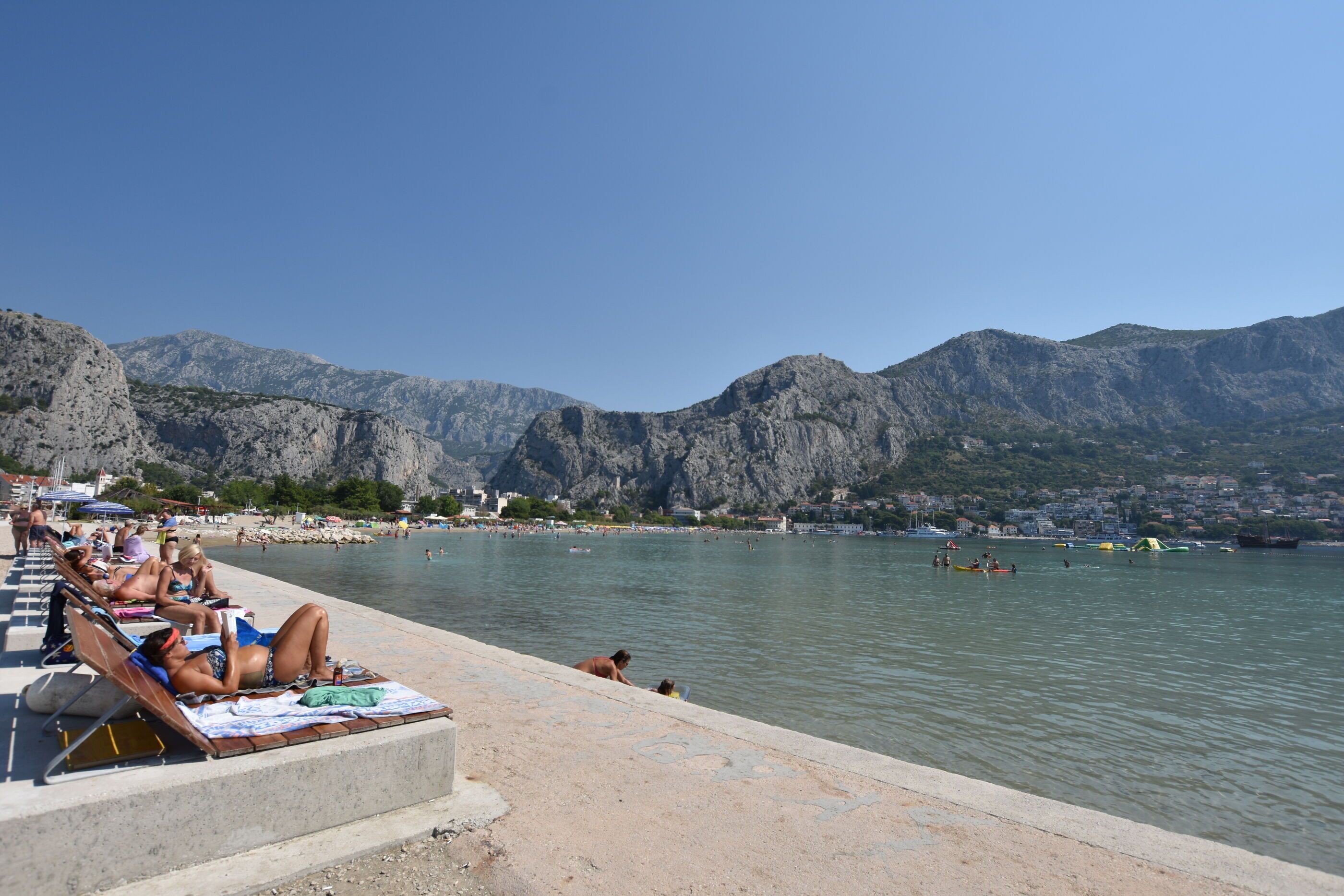 Una playa con gente tomando el sol y nadando, montañas al fondo y un cielo despejado.