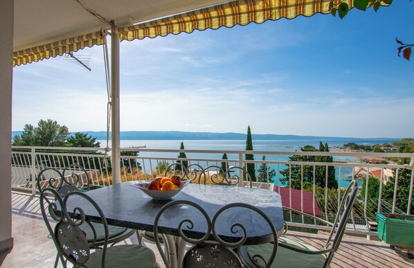 Un balcon avec une table et des chaises, un panier de fruits et une vue imprenable sur la mer et les montagnes.