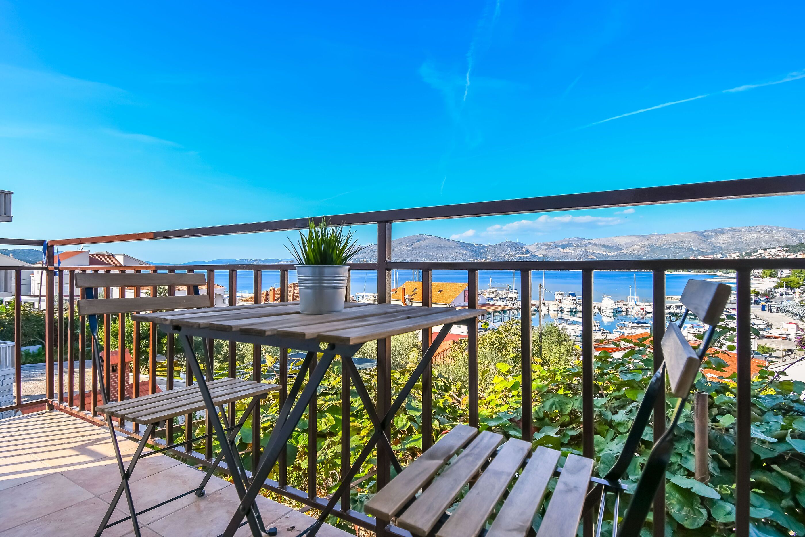 A balcony with a wooden table and chairs, a potted plant, and a view of a marina and mountains.