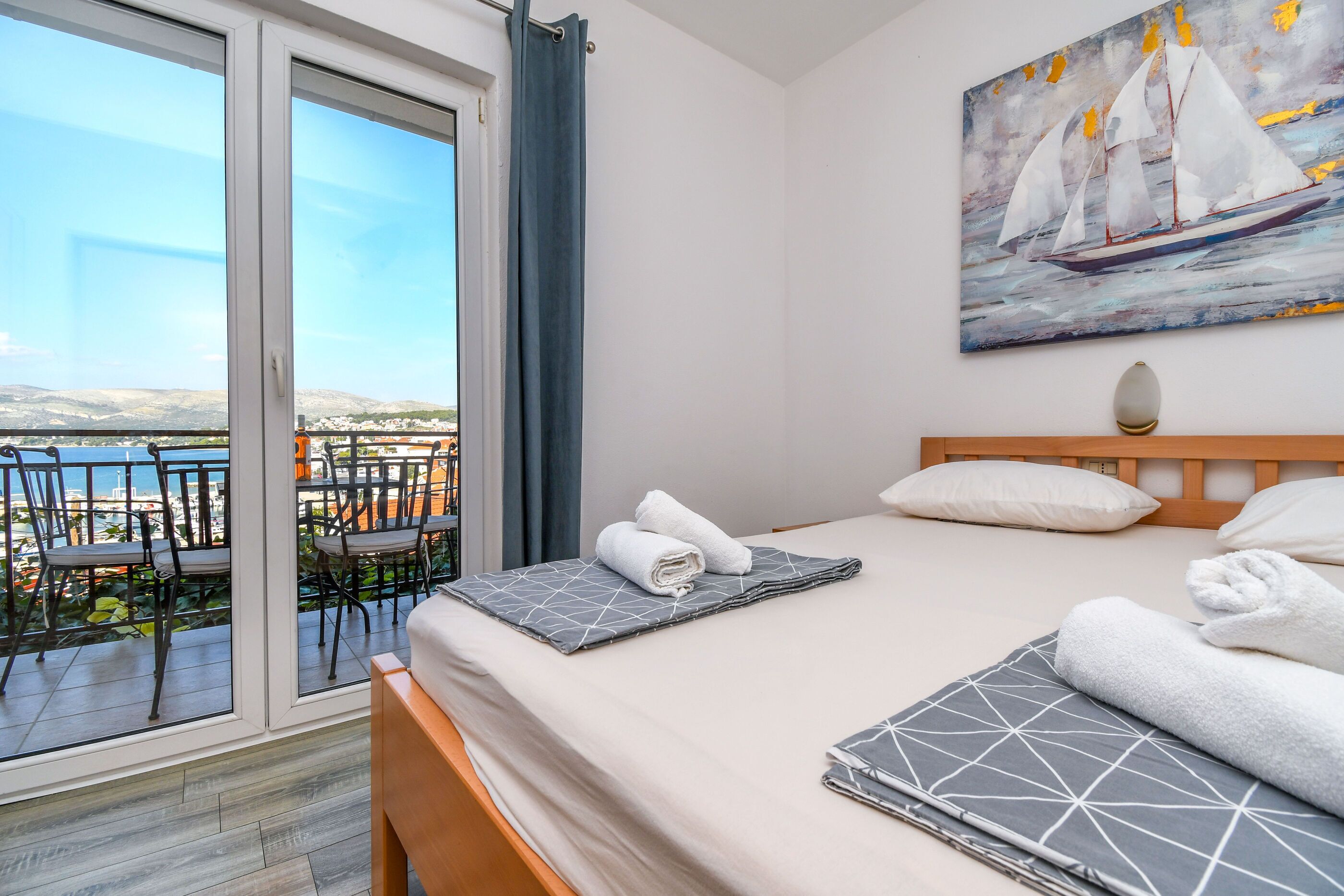 Una habitación de hotel con una cama, un cuadro de veleros, un balcón con sillas y vistas al mar.