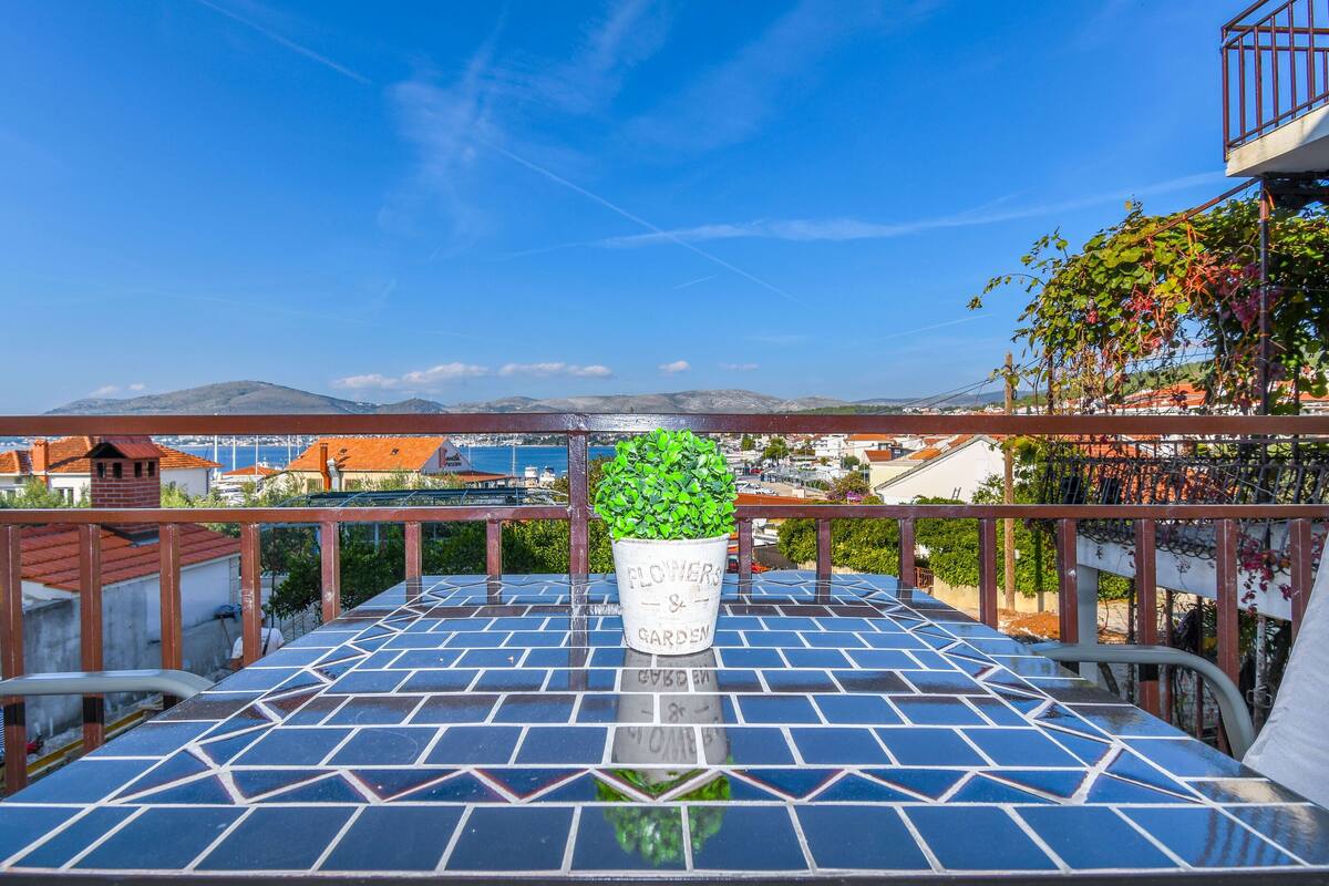 Un balcone piastrellato con una pianta in vaso etichettata 'FLOWERS & GARDEN' e vista su edifici e montagne sullo sfondo.
