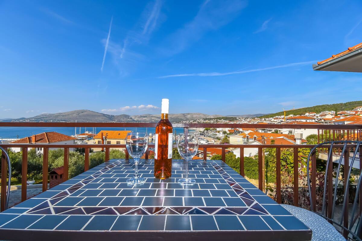 Un tavolo con una bottiglia e dei bicchieri su un balcone con vista su una cittadina costiera.