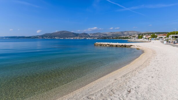 Una spiaggia costiera con acque limpide, ciottoli e una cittadina in lontananza.